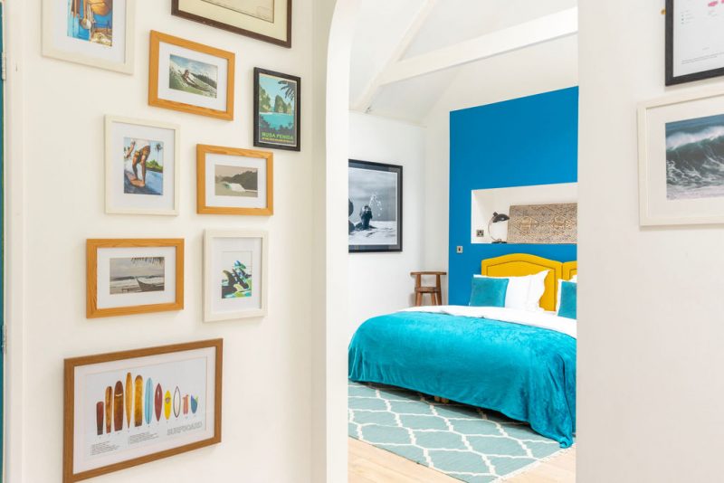Bright health retreat bedroom with a yellow headboard, turquoise bedspread, patterned rug, and framed photos on white walls.