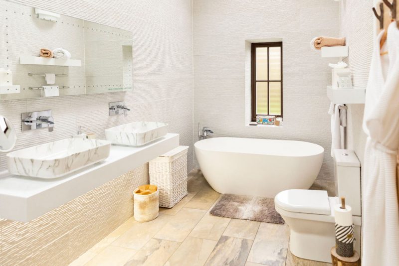 Bright luxury spa retreat like modern bathroom with a freestanding oval bathtub, dual marble sinks, toilet, and beige tiled floor.
