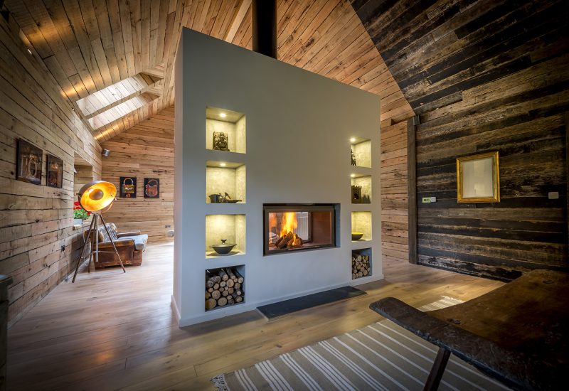 Luxury rustic retreat living room with a central modern fireplace featuring built-in illuminated shelves and stacked firewood, surrounded by wooden walls and floors.