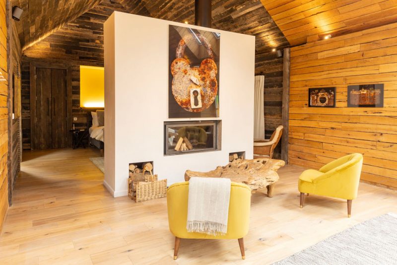 retreat cabin interior with wooden walls, a white fireplace featuring art of a rusty lock, two yellow armchairs, and a rustic wooden coffee table.