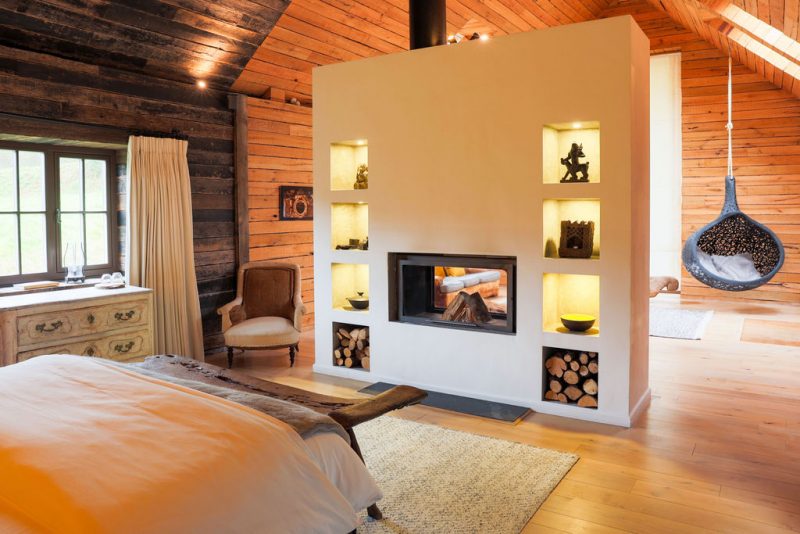 Wellness Retreat styled bedroom in a wooden cabin featuring a modern fireplace with built-in shelves, cushioned armchair, rustic dresser, hanging wicker chair, and skylight.