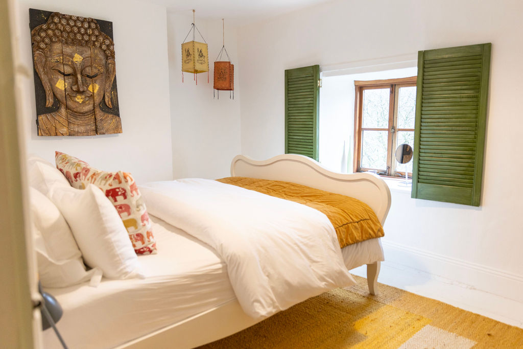 Bright bedroom in a health retreat with white walls, a white bed with elephant-print pillows, a mustard-colored quilt, Buddha wall art, and hanging lanterns.