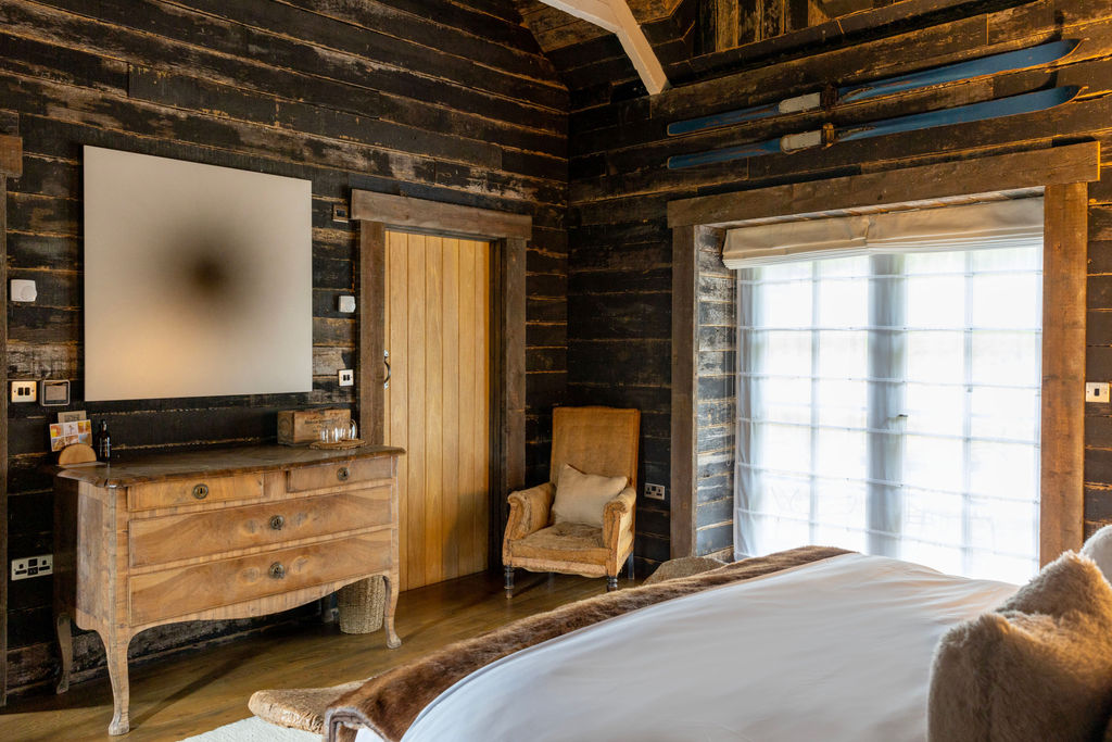 English UK health retreat bedroom with a patterned bed, yellow striped blanket, two yellow pillows, wooden bench, rustic walls, and framed artwork.