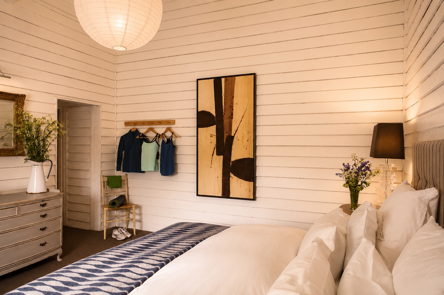 Double bed with white pillows and a fur blanket against an olive green wall with a wooden nightstand and a decorative lamp.