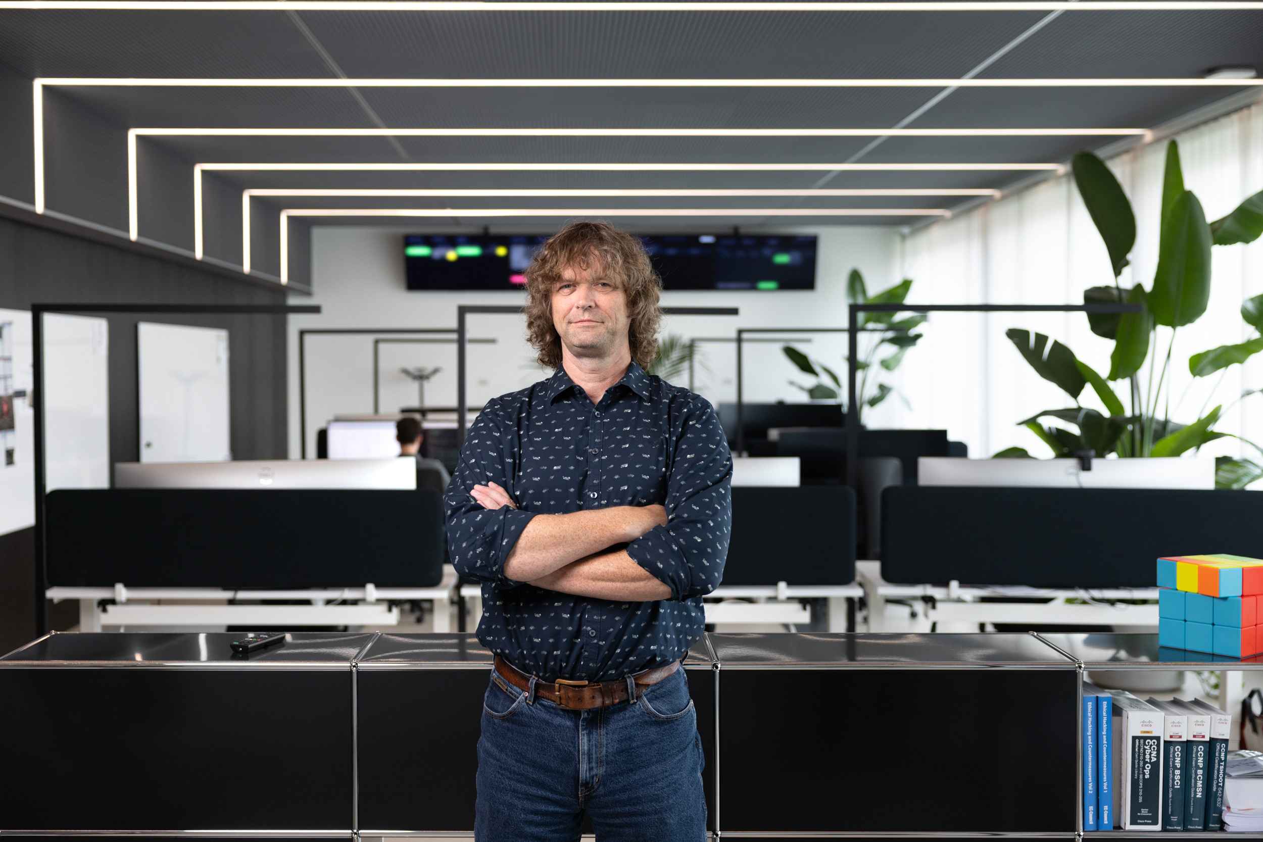 Ein Mann mit verschränkten Armen steht in einem modernen Büro mit Computerarbeitsplätzen und großen Grünpflanzen.