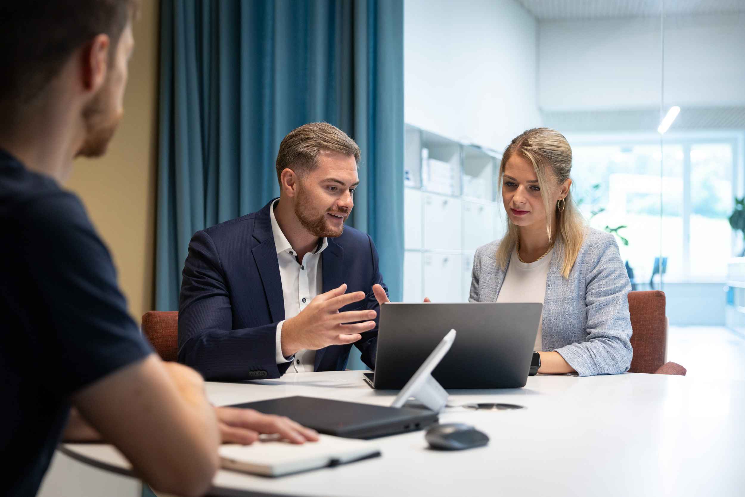 Zwei Geschäftsleute diskutieren vor einem Laptop, während eine dritte Person im Vordergrund zuhört.