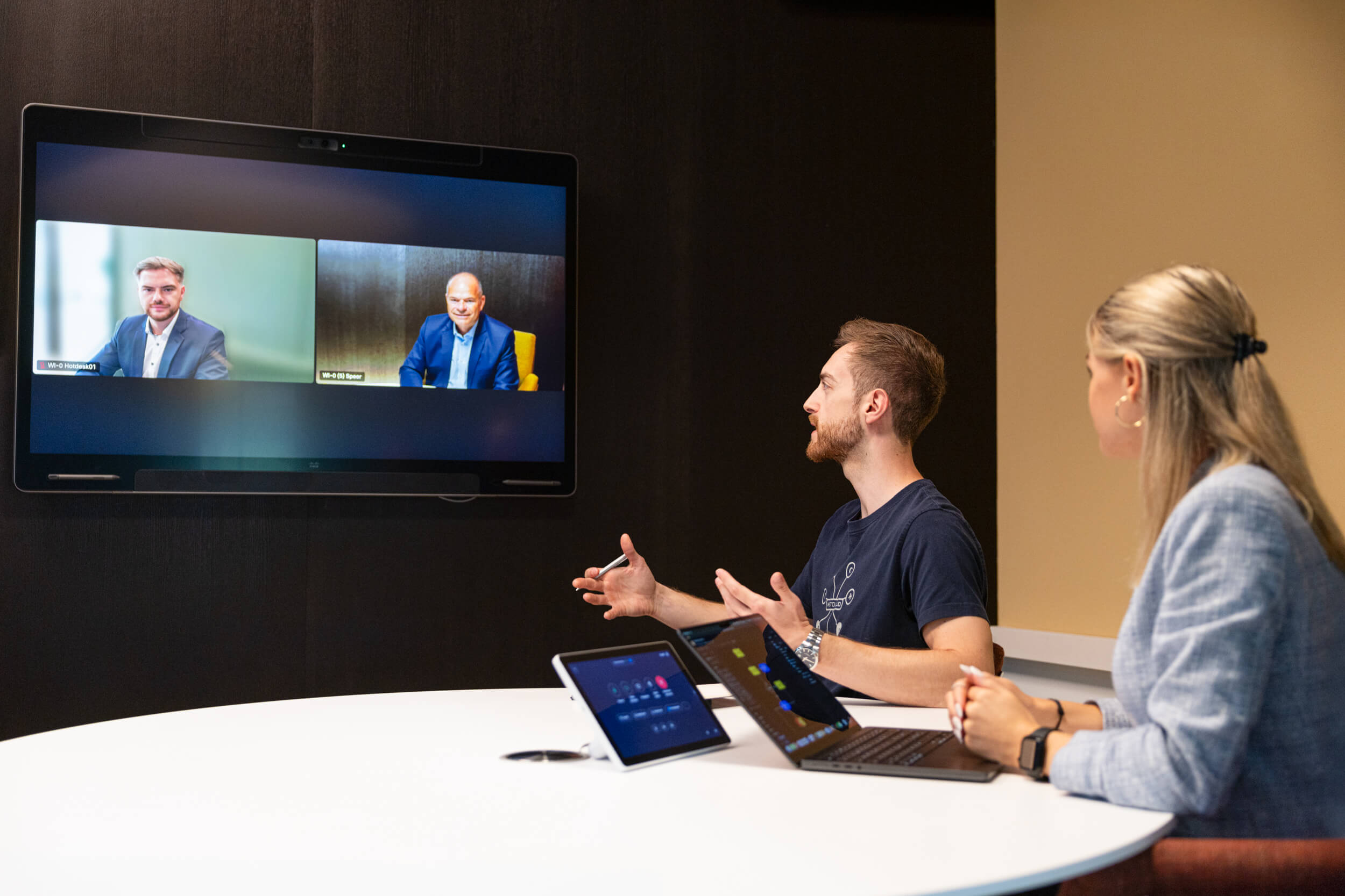 Zwei Personen im Büro sitzen am runden Tisch und sprechen in einer Videokonferenz mit zwei Männern auf einem Bildschirm.