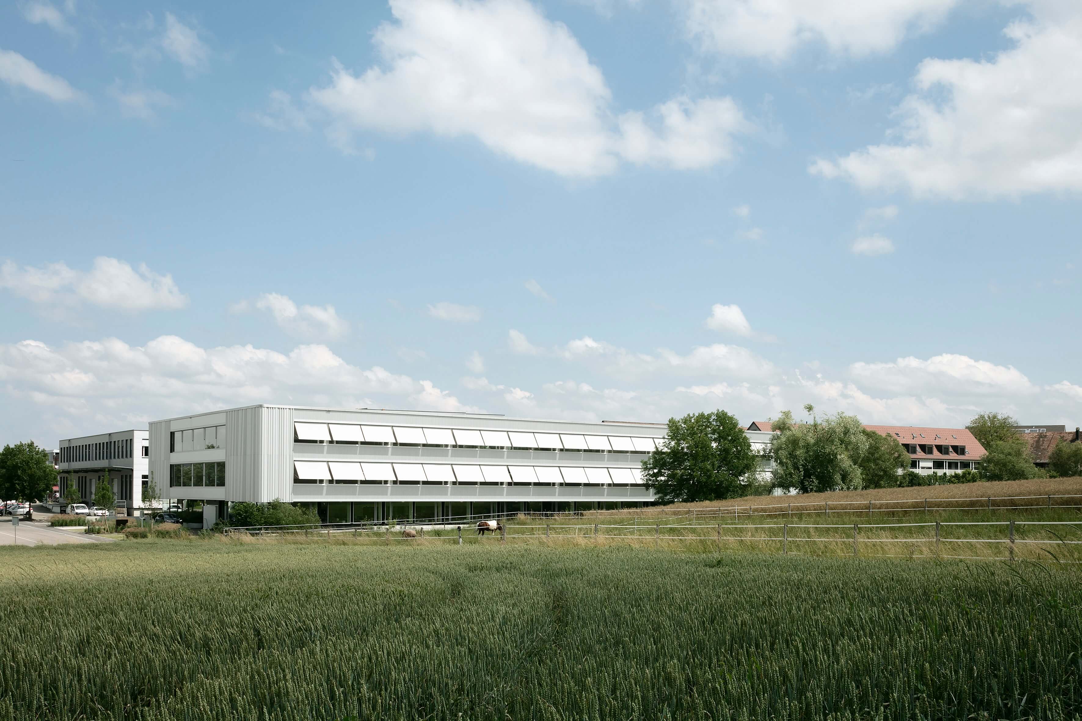 Modernes Bürogebäude mit weißen Fassadenfronten, umgeben von einem grünen Feld und Bäumen unter blauem Himmel mit Wolken.