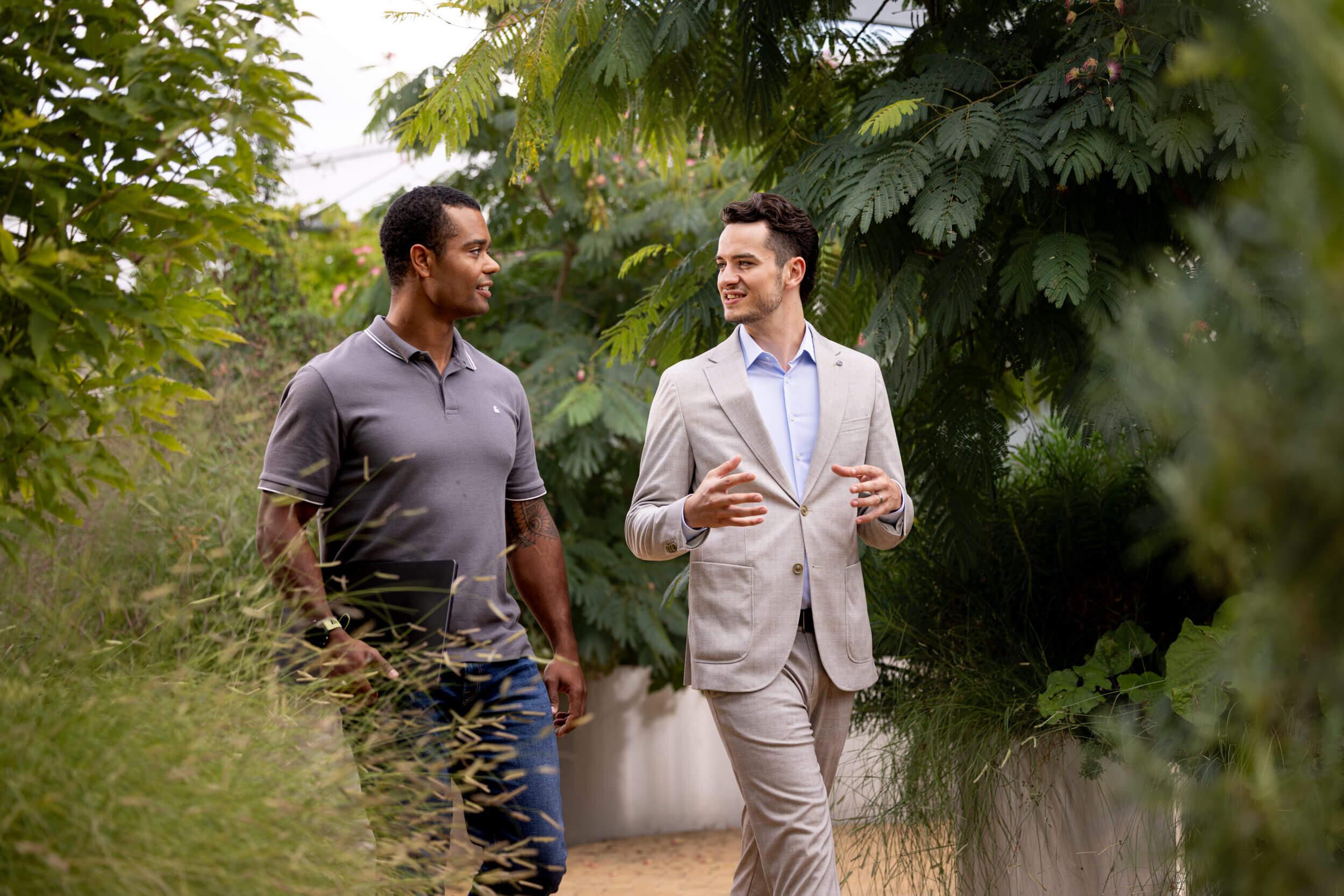 Zwei Männer in einem grünen Garten, einer trägt ein graues Poloshirt und Jeans, der andere einen hellen Anzug, beide im Gespräch.