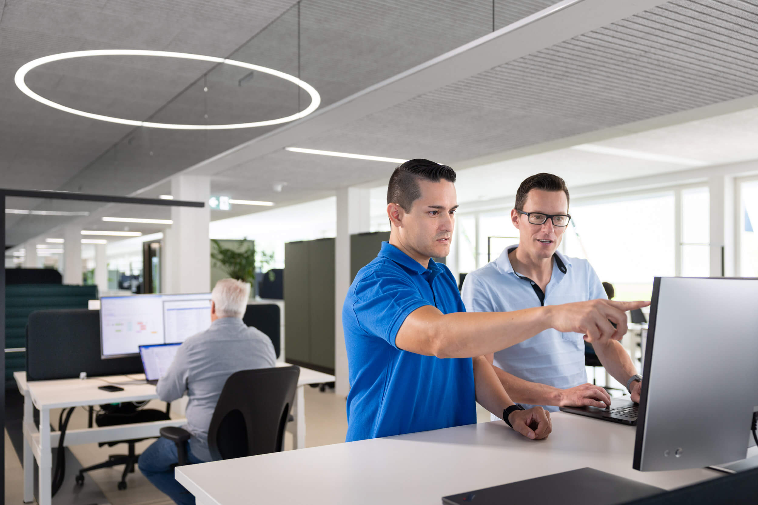 Zwei Männer stehen an einem Stehpult im modernen Büro, einer zeigt auf den Computermonitor, im Hintergrund arbeitet ein älterer Mann am Schreibtisch.