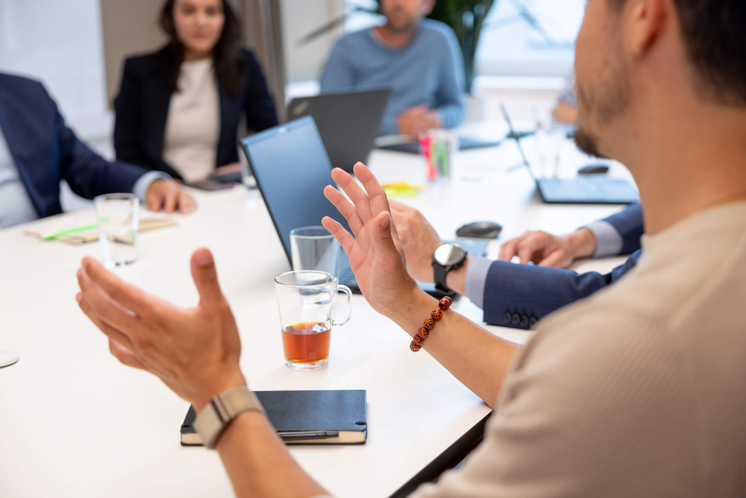 Person gestikuliert während einer Besprechung mit mehreren Personen an einem Besprechungstisch.