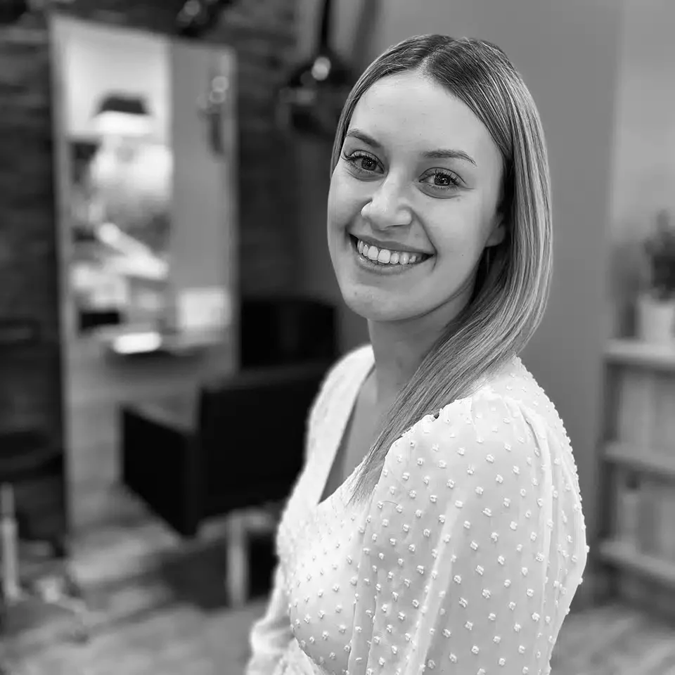 Portrait en noir et blanc d'une femme souriante aux cheveux longs, portant un haut texturé, dans un intérieur flou.