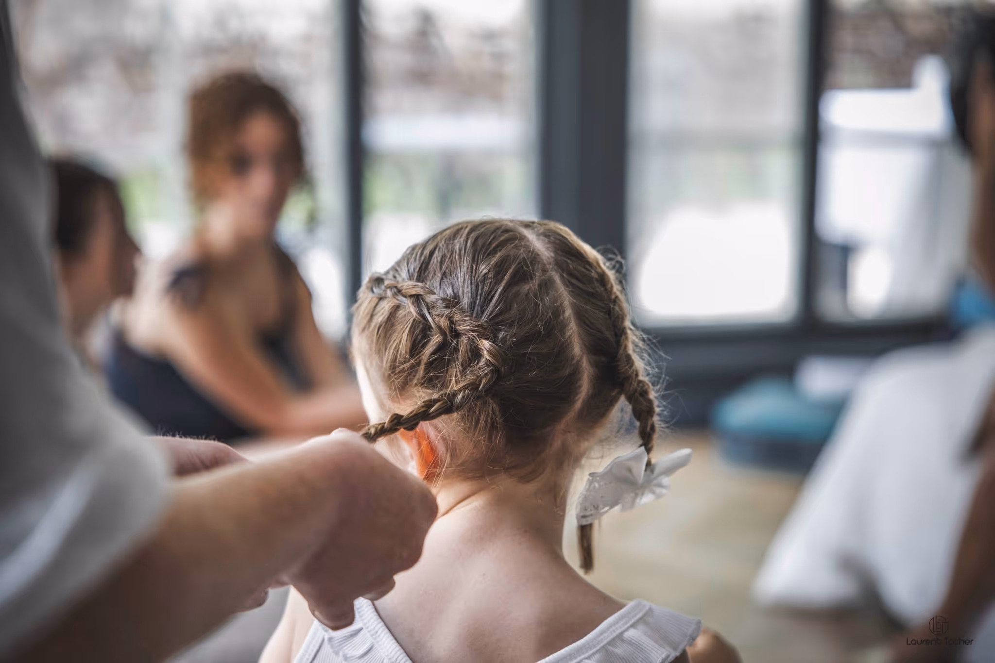 Une enfant vue de dos avec des tresses dans les cheveux, une personne stylise ses cheveux avec un ruban blanc.