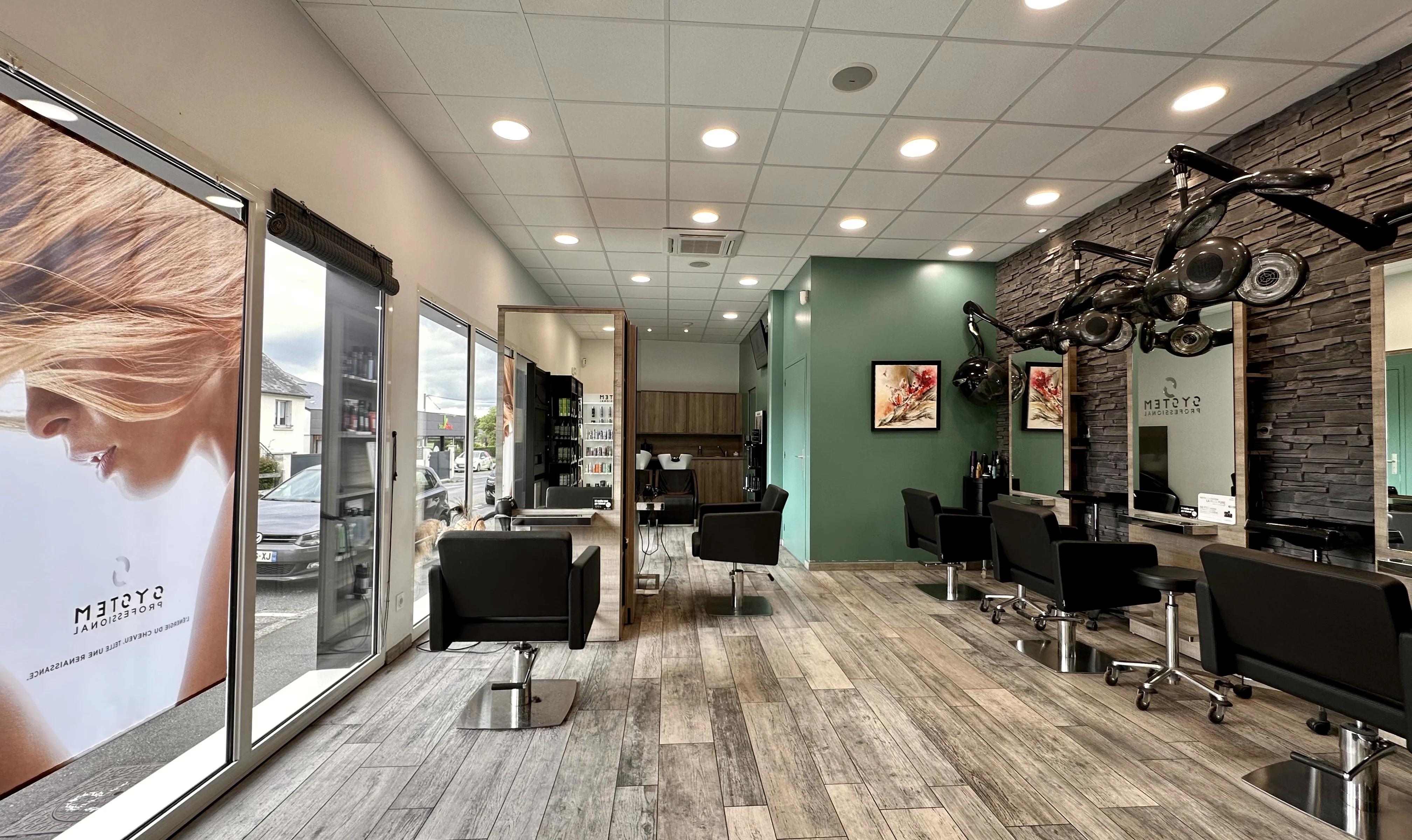 Salon de coiffure moderne avec fauteuils noirs, grandes fenêtres sur la gauche et murs décorés de pierres et tableaux.