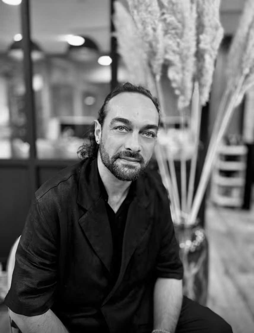 Portrait en noir et blanc d'un homme aux cheveux longs attachés, barbe, portant une veste noire, assis avec un grand vase de plantes en arrière-plan flou.