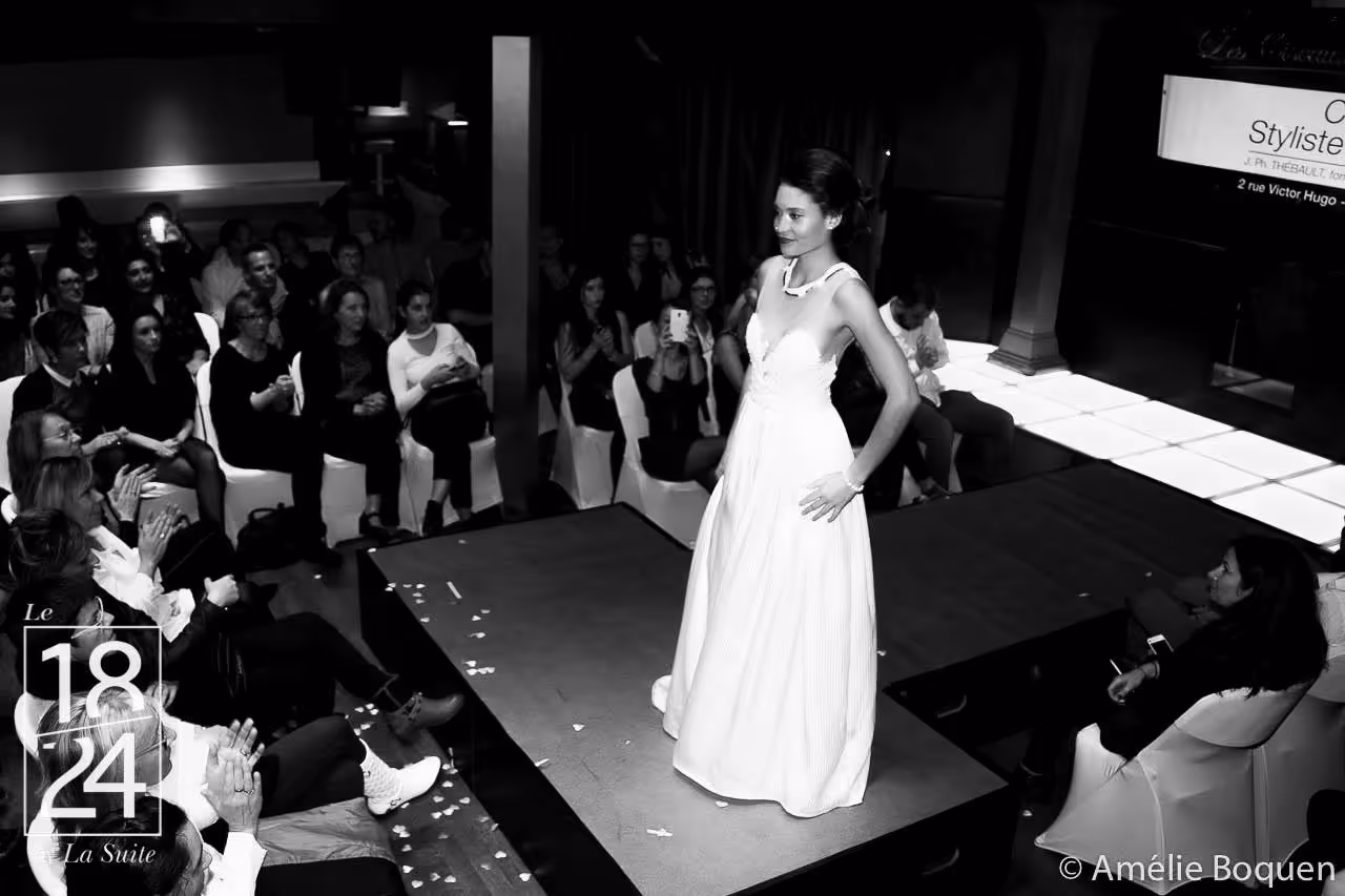 Une mannequin sur un podium lors d'un défilé de mode, avec un public assis autour qui l'applaudit.