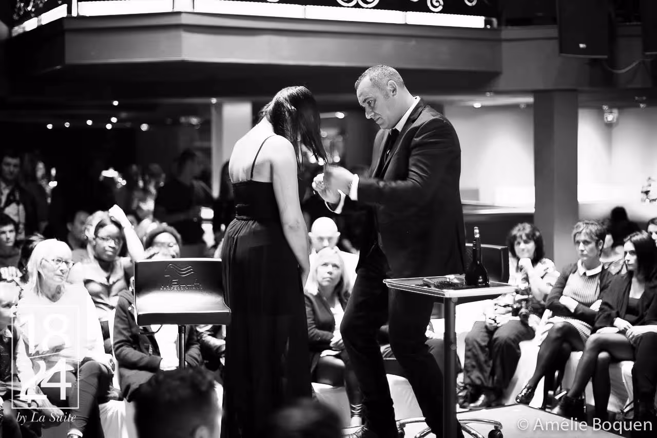 Un homme en costume effectue une démonstration de coiffure sur une femme en robe noire devant un public attentif.