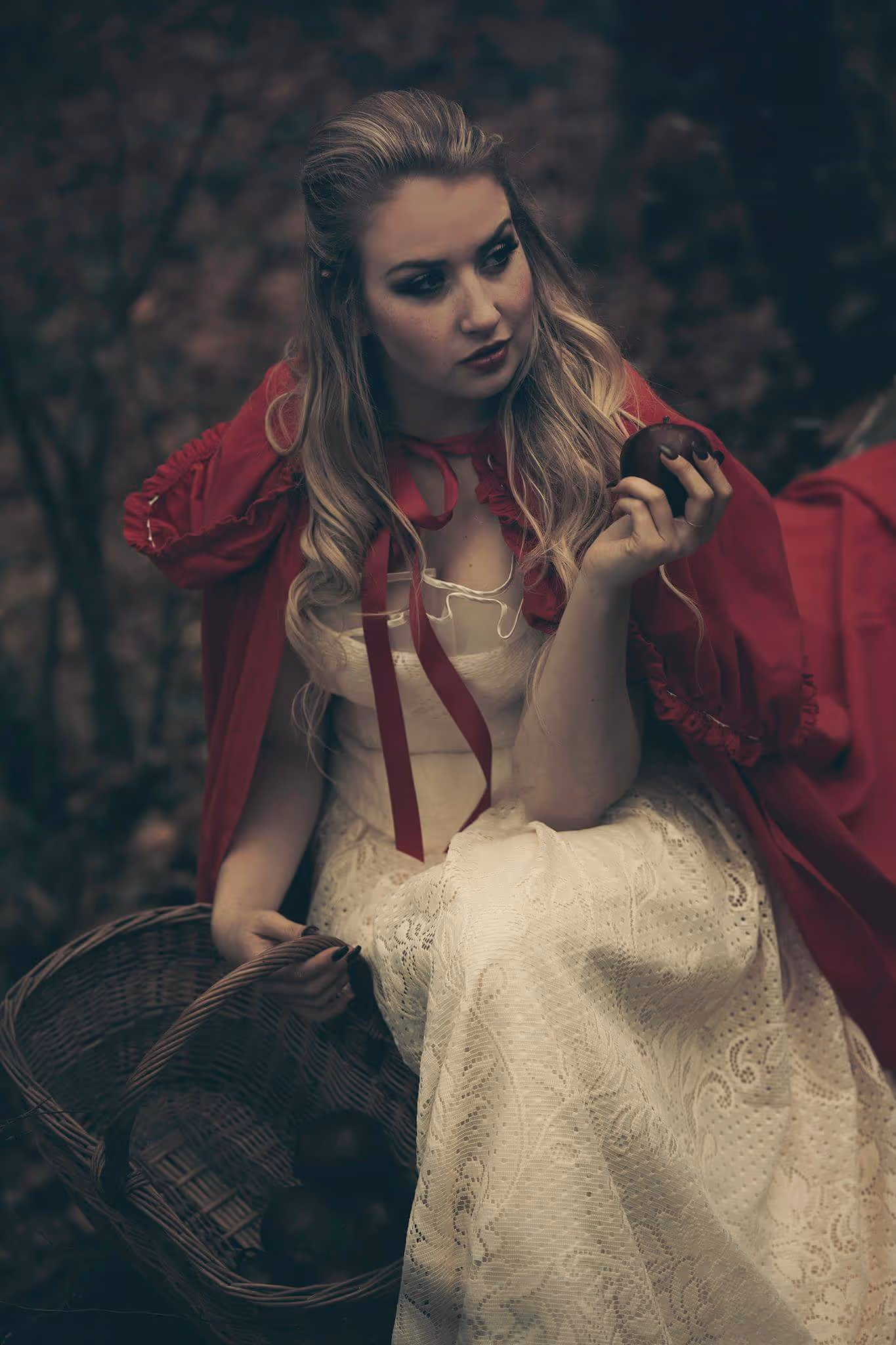 Femme blonde en robe blanche en dentelle et cape rouge tenant une pomme dans une main et un panier en osier dans l'autre, assise en forêt.