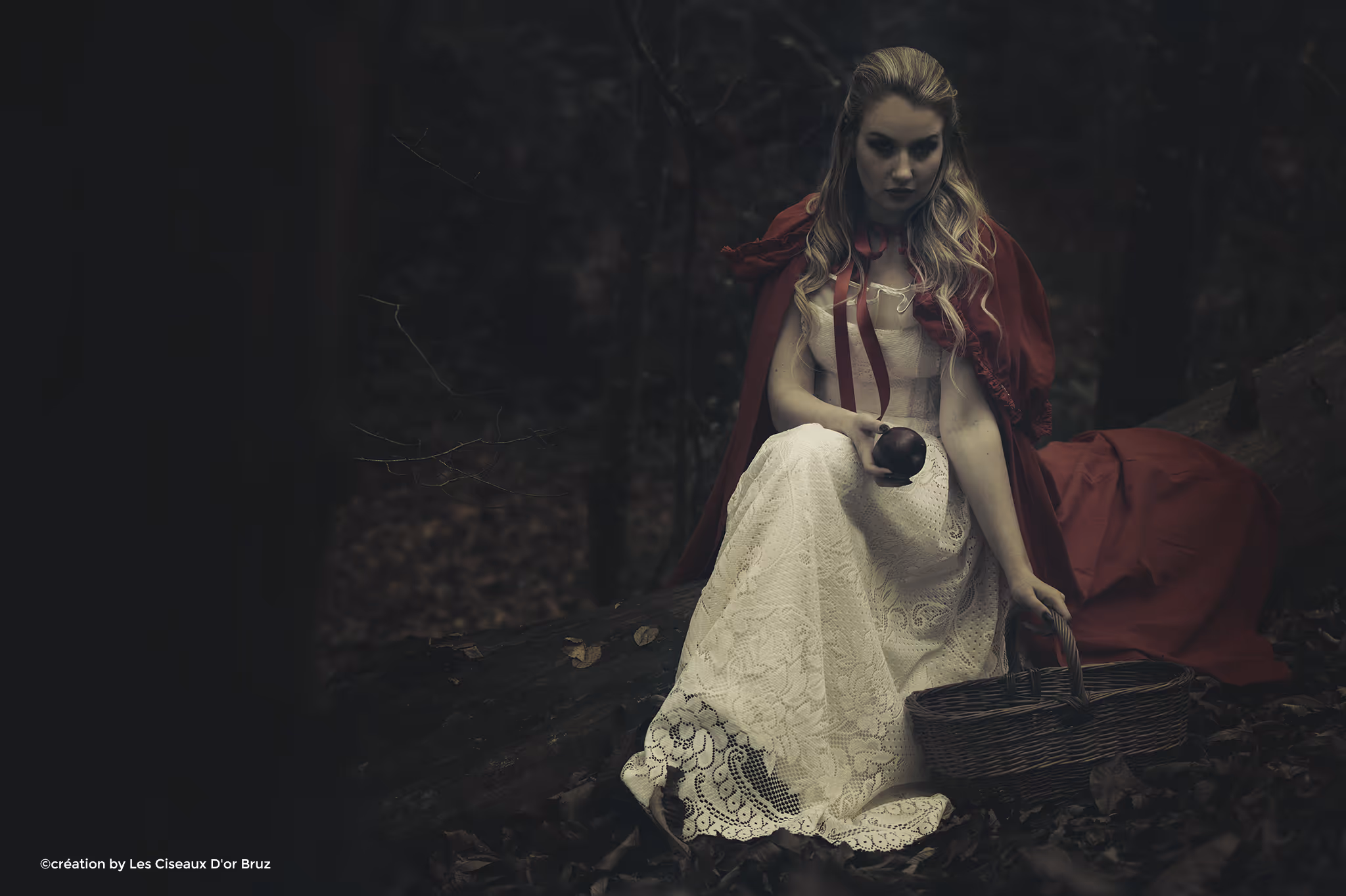 Une femme blonde en robe blanche et cape rouge assise sur une souche avec un panier et une pomme dans une forêt sombre.