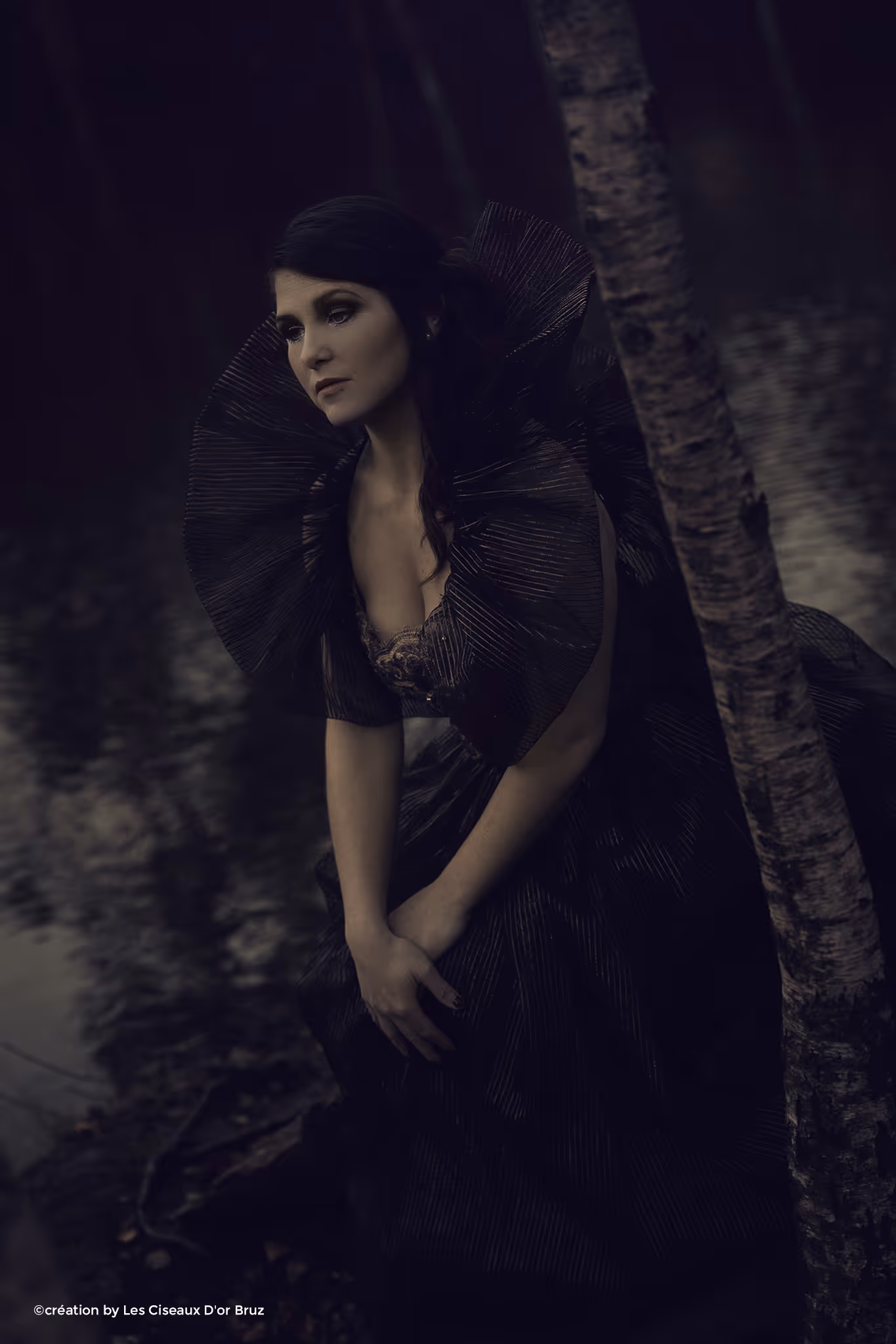 Femme vêtue d'une robe noire élégante au col large en tissu plissé, assise près d'un arbre au bord de l'eau, regard pensif.