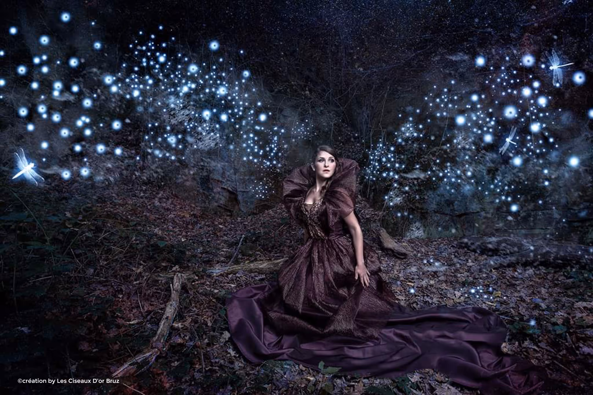 Femme en robe violette longue assise dans une forêt sombre entourée de lucioles lumineuses et de libellules éclatantes.