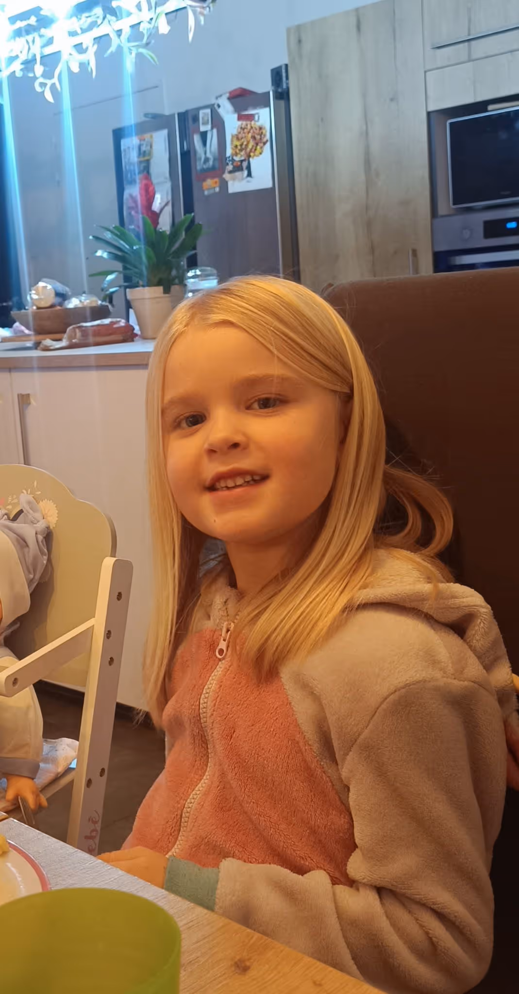 Jeune fille blonde souriante portant une veste douce, assise à une table dans une cuisine moderne.