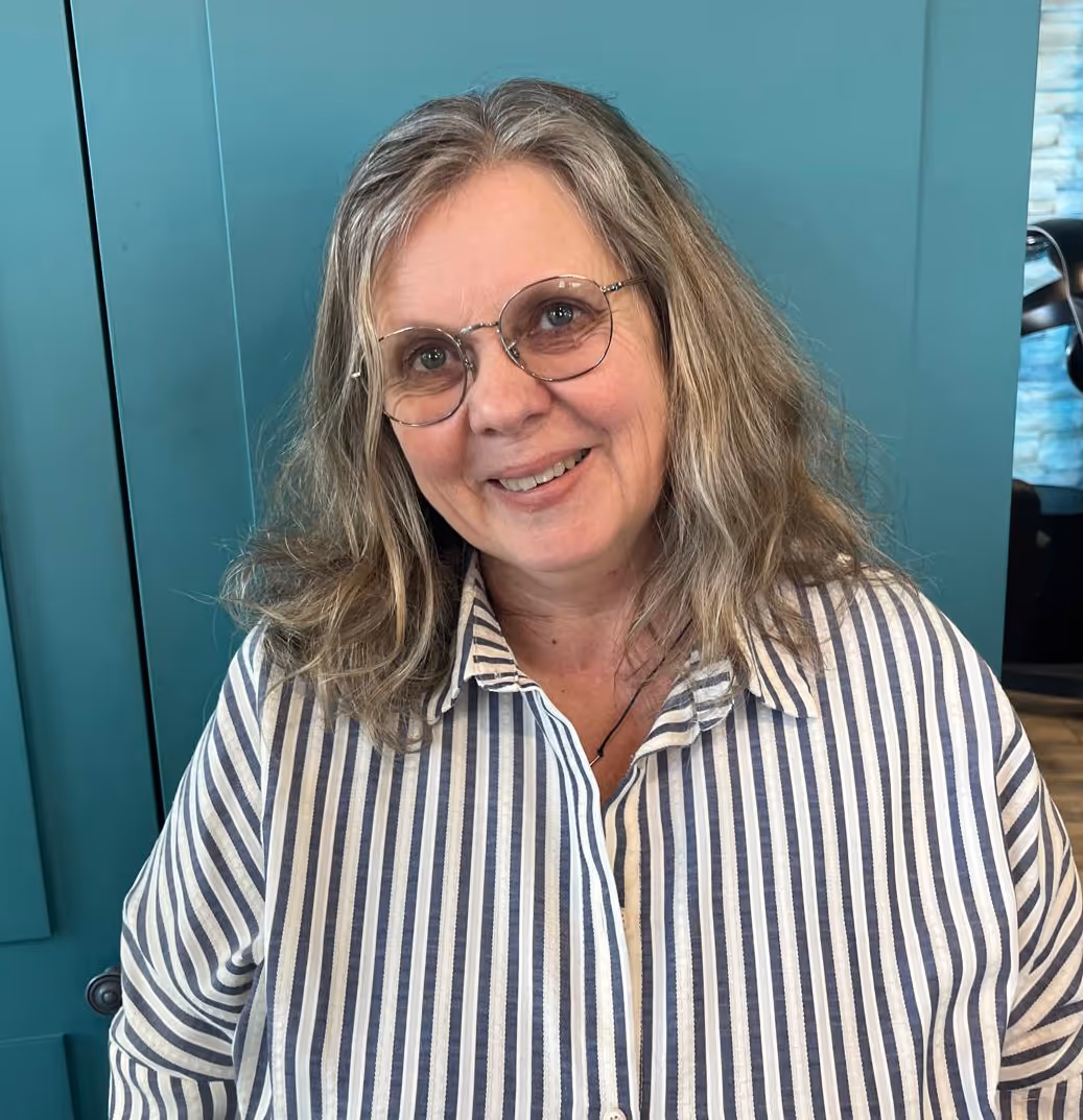 Femme souriante portant une chemise à rayures verticales blanches et bleues et des lunettes, devant un mur bleu.