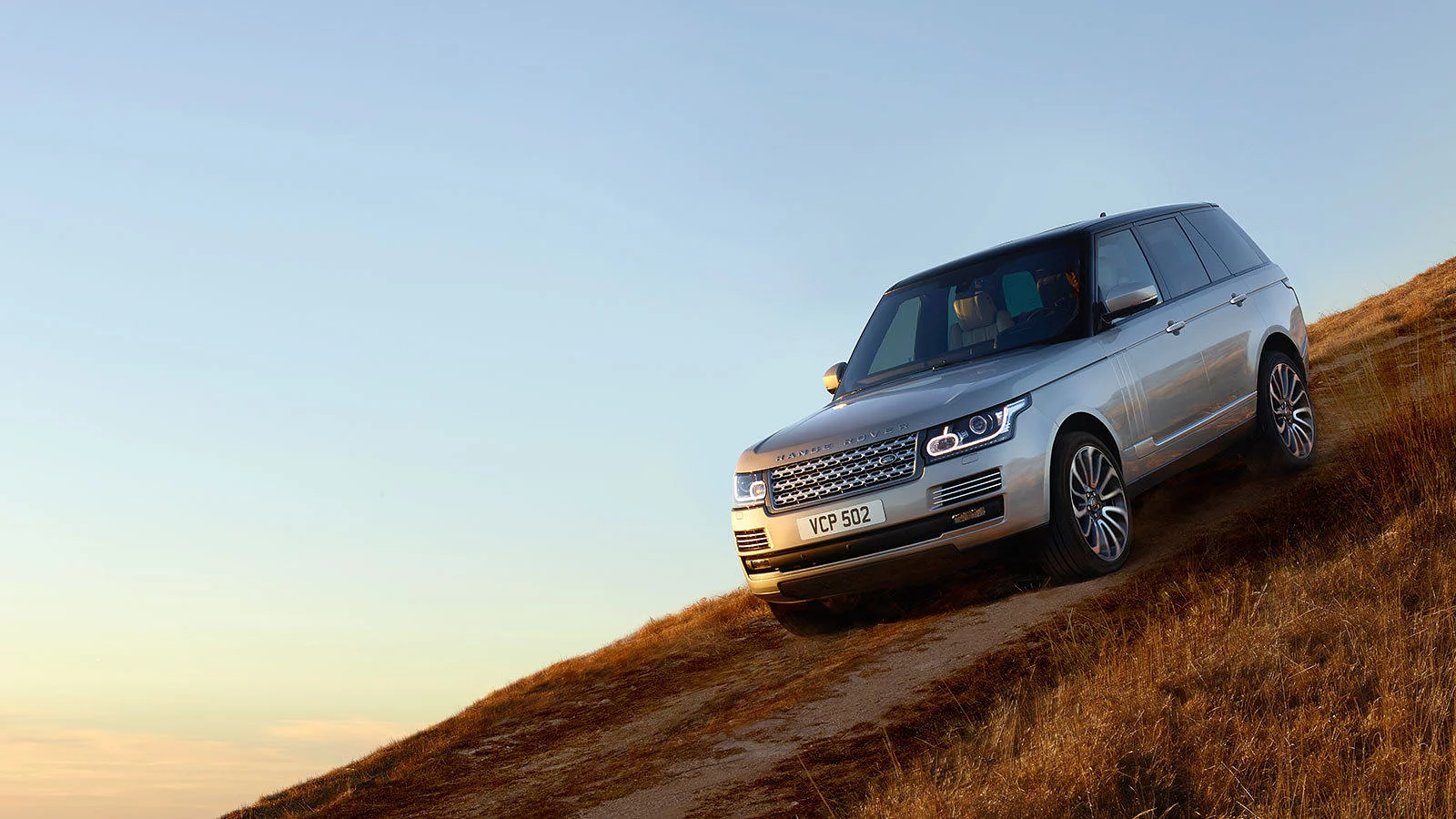Silver Range Rover driving down a steep grassy hill under a clear sky at sunset.