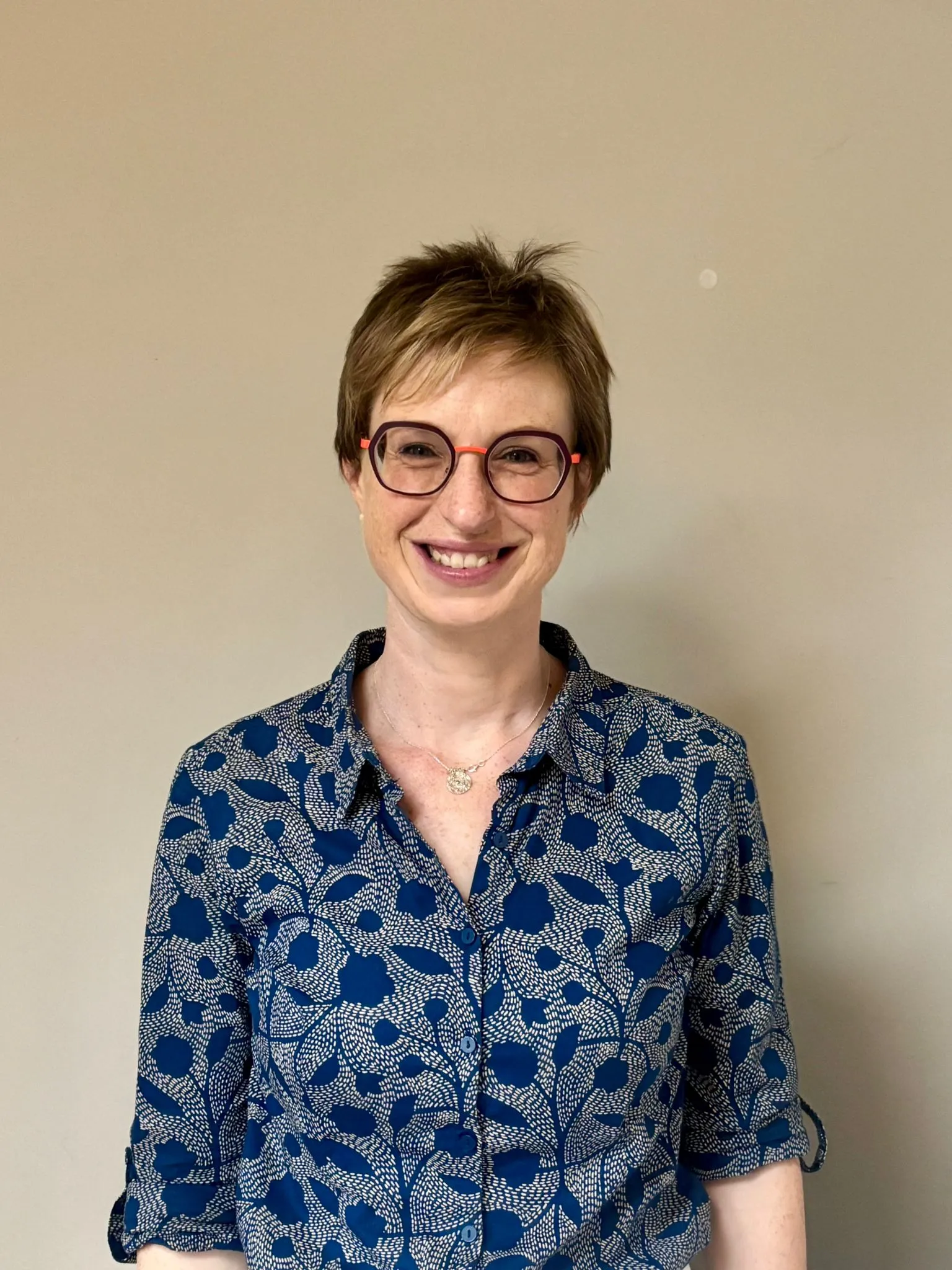 Smiling person in blue patterned shirt and glasses against neutral background