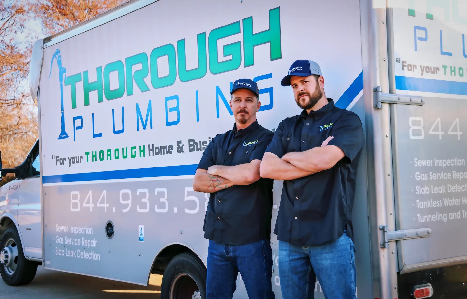 Two men in black Thorough Plumbing uniforms and caps standing with arms crossed in front of a company truck.