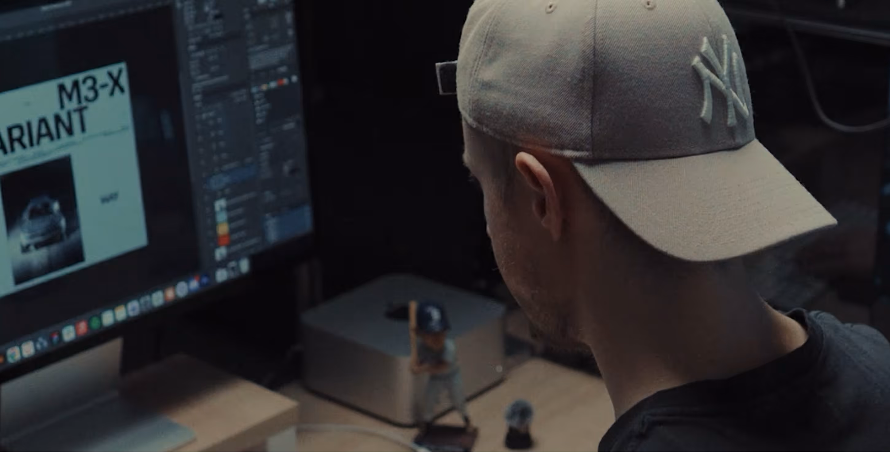 Man wearing a beige New York Yankees cap working at a computer desk with a large monitor displaying a design project.