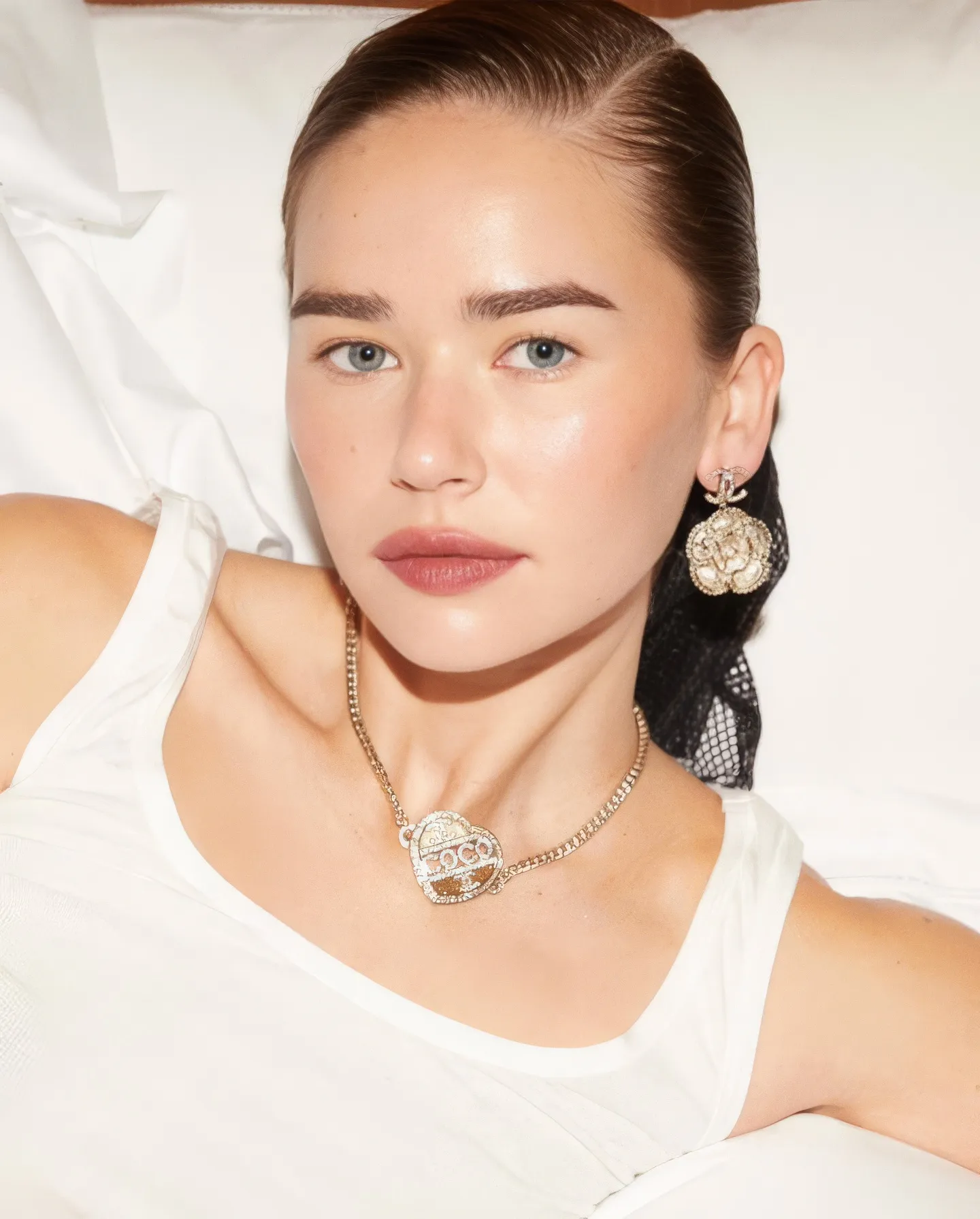 Close-up portrait of a woman with slicked-back hair wearing white and gold flower earrings and a heart necklace.