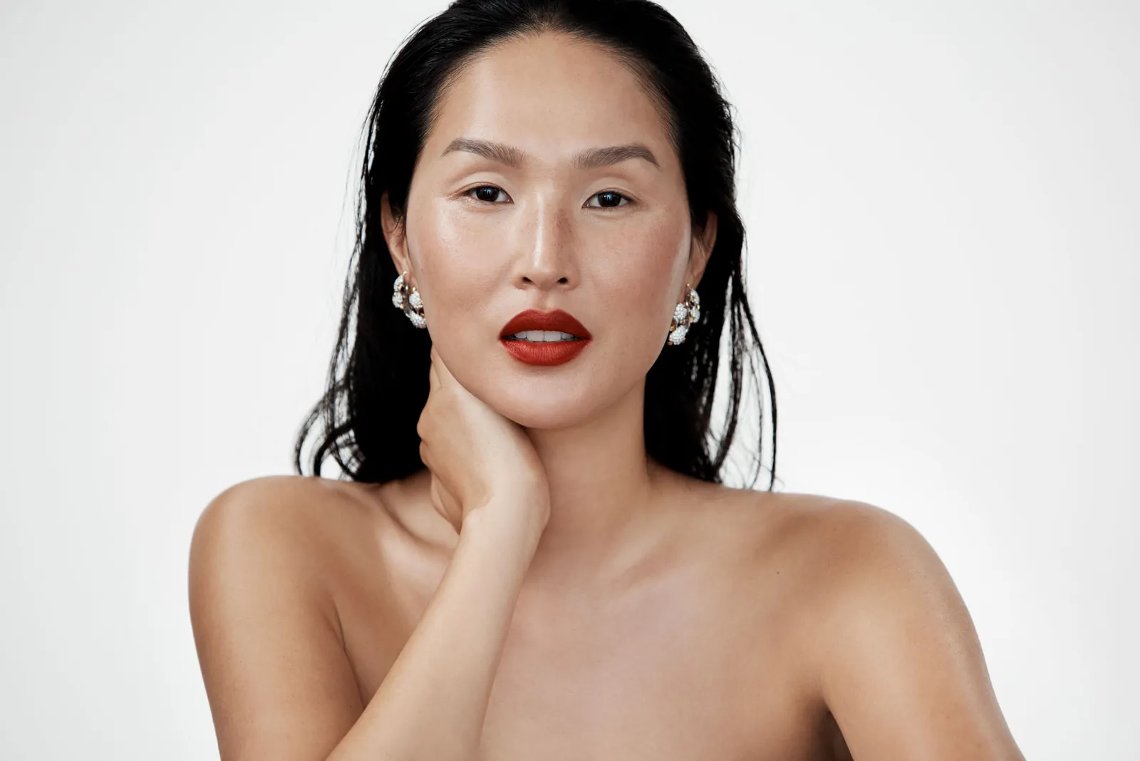 A woman with glowing skin, red lipstick and pearl earrings, her hand at her neck.