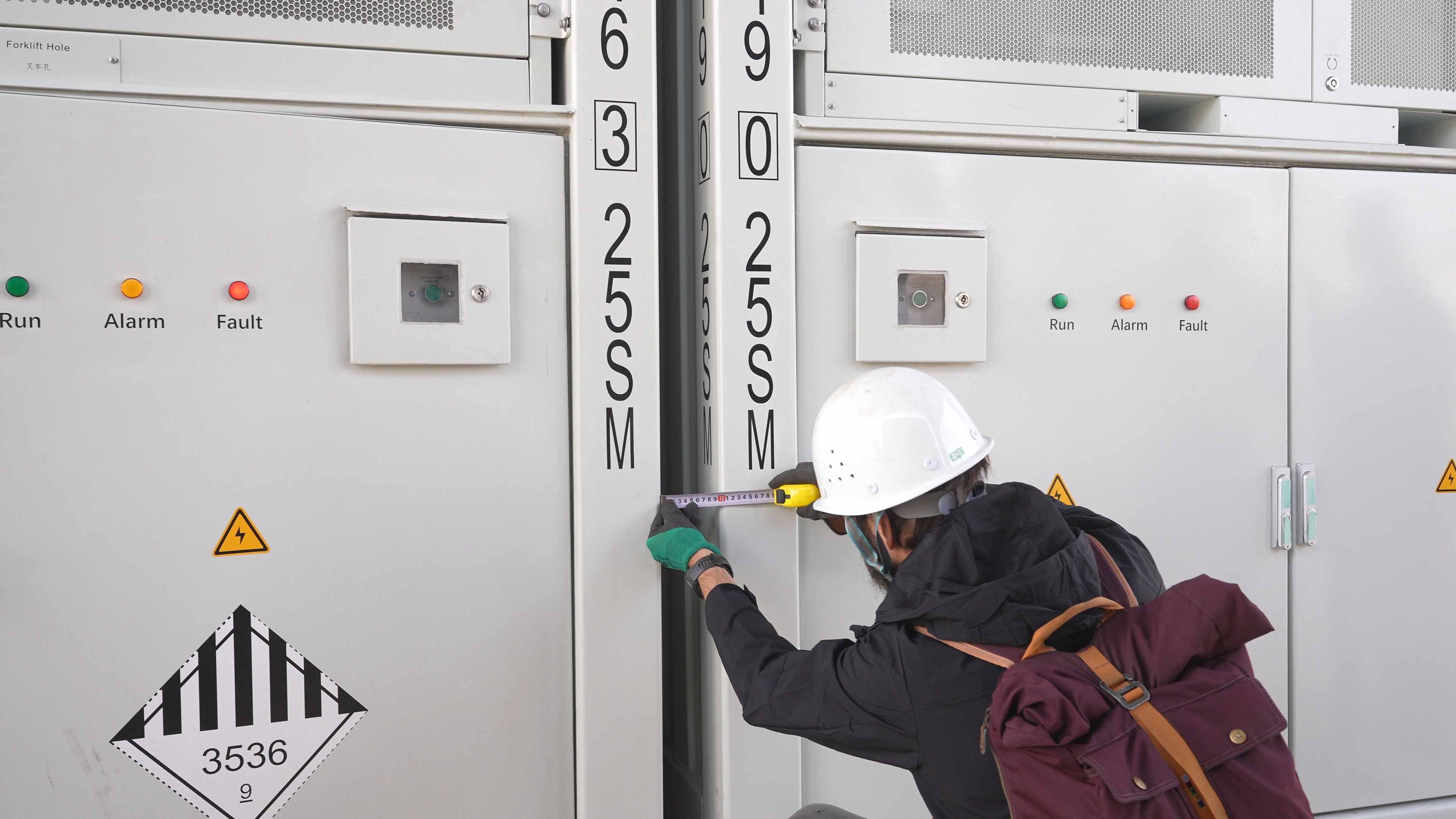 Worker wearing a white helmet and backpack measuring the gap between electrical panels labeled 6300 25SM using a tape measure.