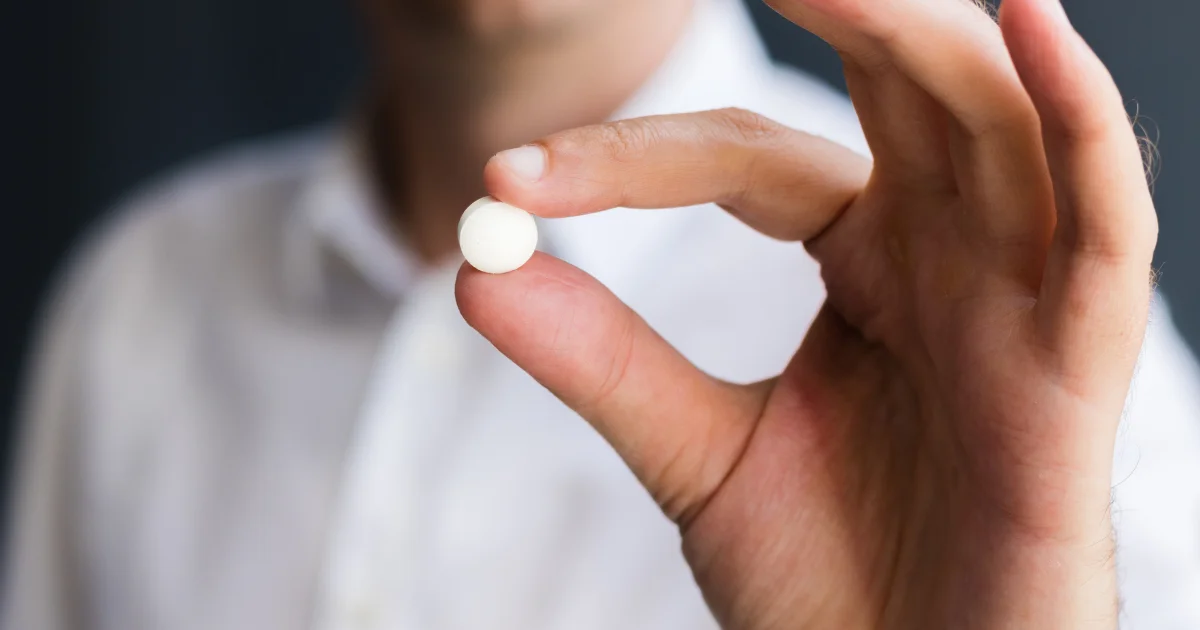 Man in a white shirt holding a single white oral GLP-1 weight loss pill between finger and thumb