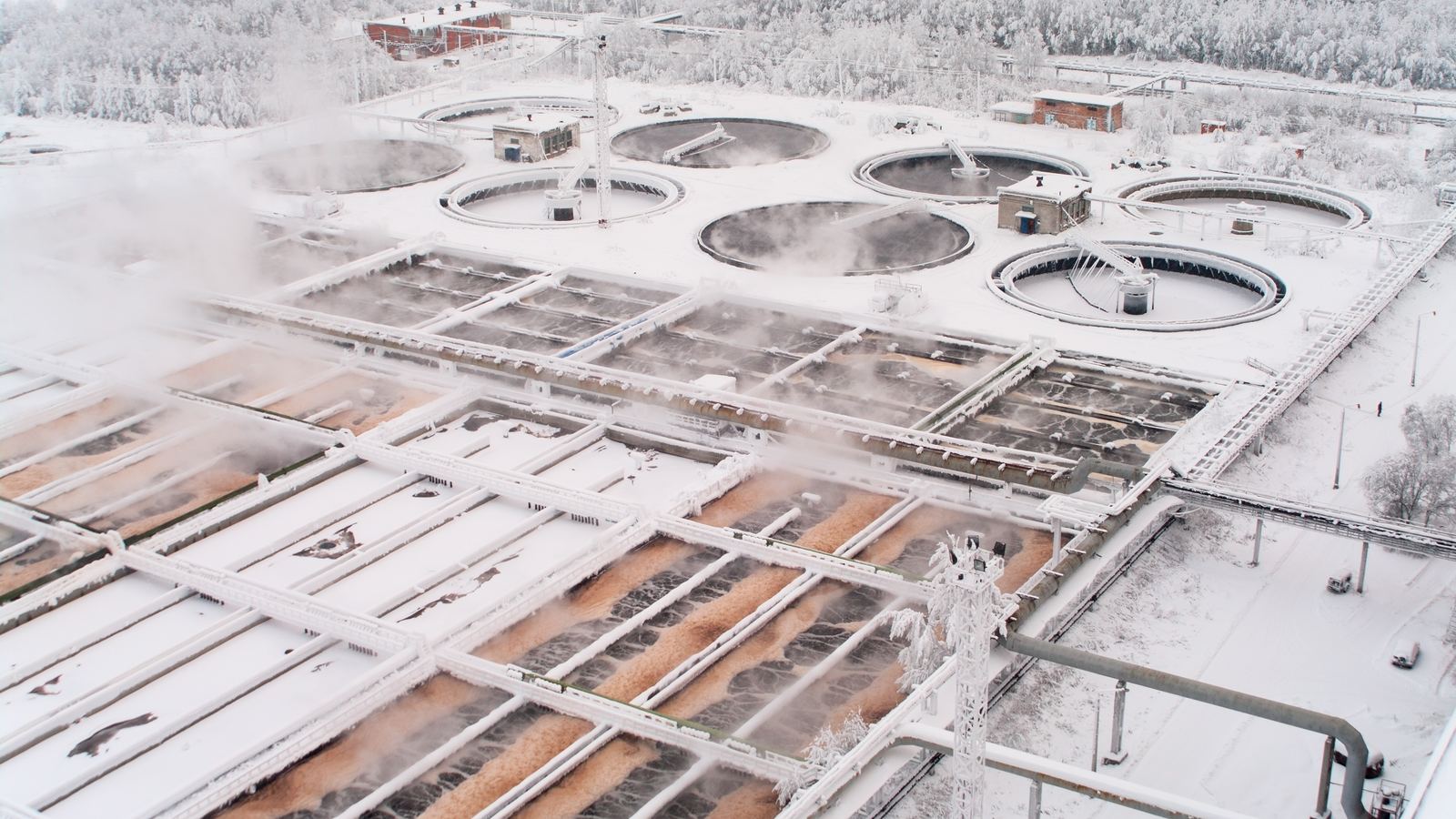 Aerial view of a wastewater treatment plant in winter, clarifiers and aeration basins steaming under snow