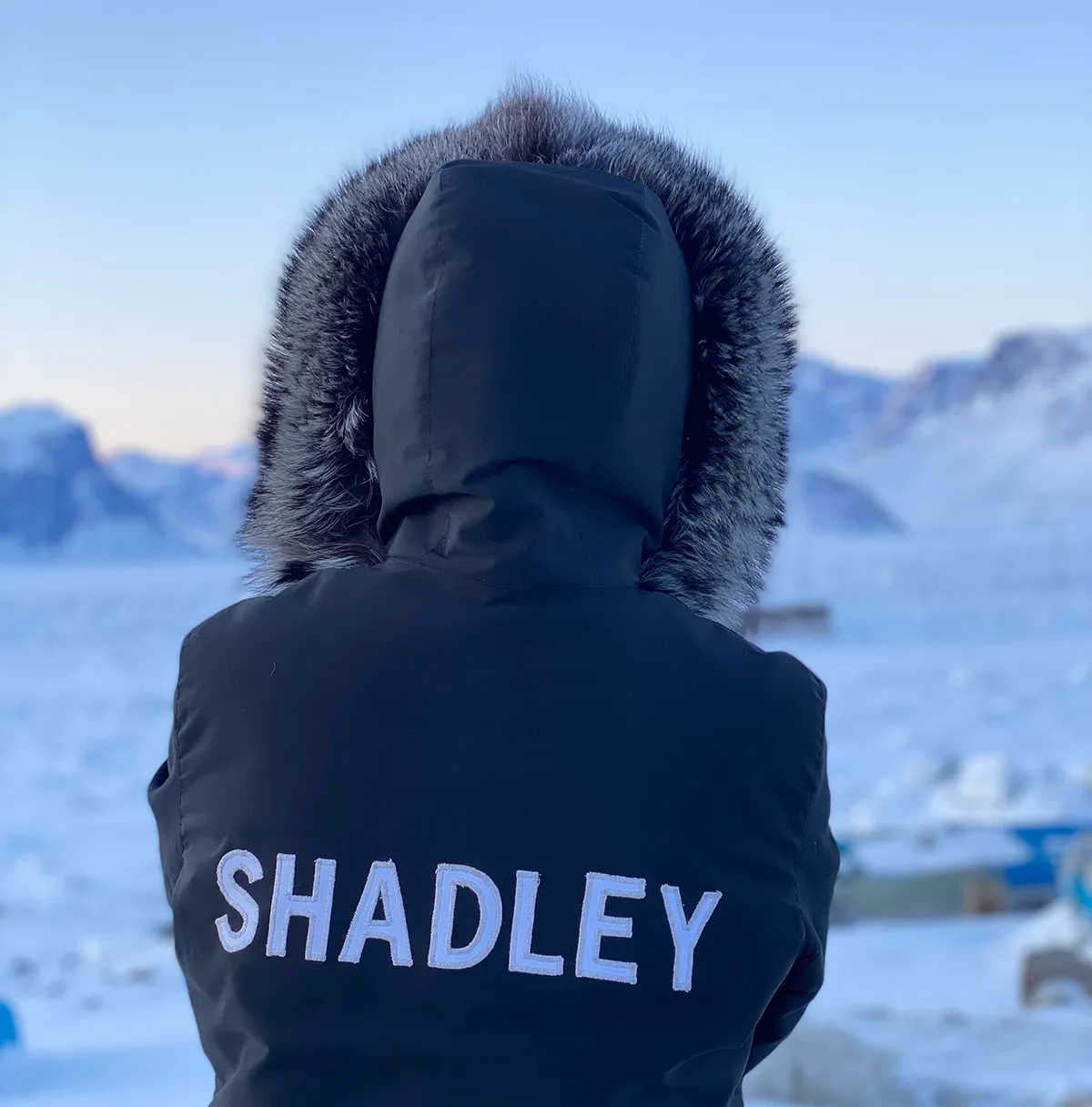 person facing away from the lens into the snowy surroundings. The person is wearing a winter parka with the fur-lined hood up and a large 'Shadley' word on the back of the parka.
