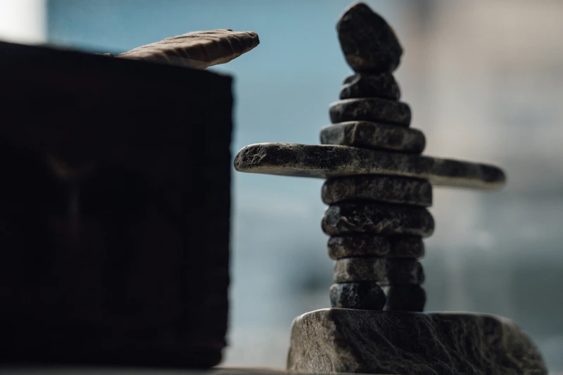 Small stacked stone sculpture resembling a human figure with outstretched arms on a flat base, set against a blurred background.