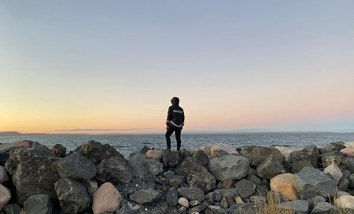 Lauren Shadley standing on a bank of large rocks facing out towards the water with a beautiful horizon.
