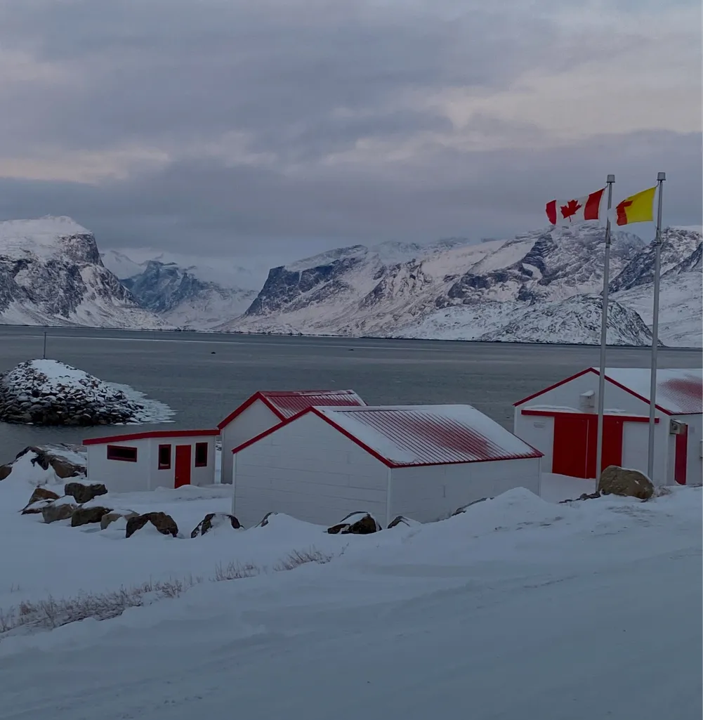 Snow-covered landscape with small white buildings featuring red roofs and doors near a body of water, with snow-covered mountains in the background and three flags on poles, including the Canadian flag.