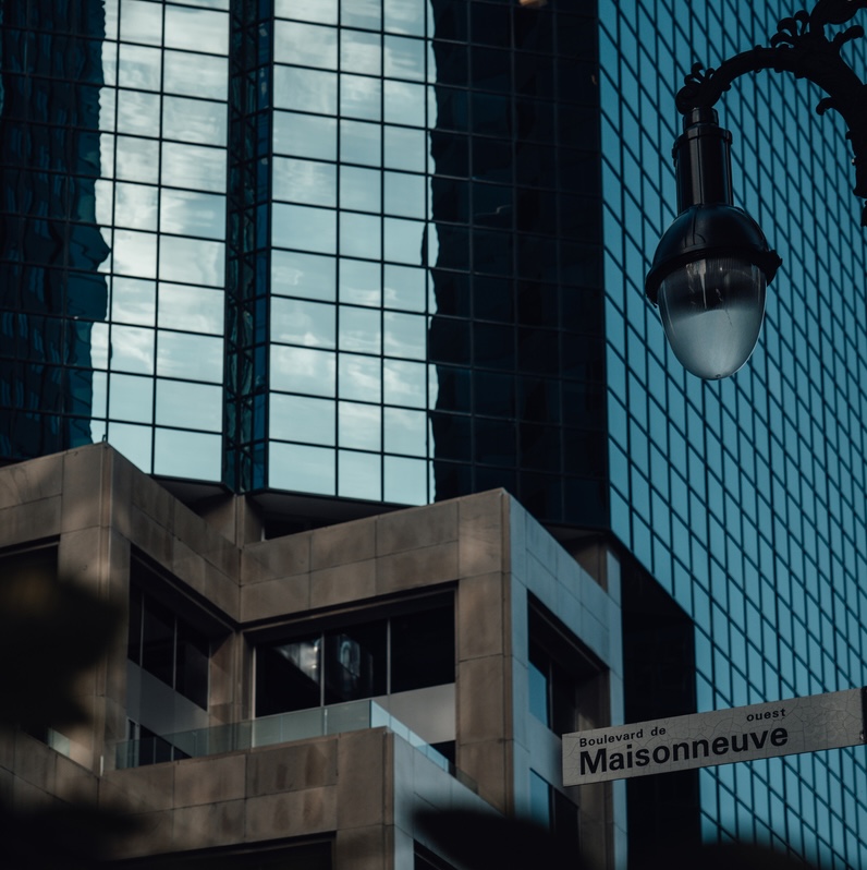 Modern office buildings with glass facades reflecting the sky and a streetlamp on the right side, with a street sign reading 'Boulevard de Maisonneuve Ouest' in the lower right corner.