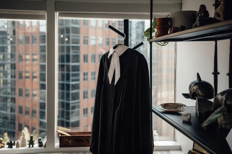 Black legal robe with white neckband hanging on a coat rack near a window with a cityscape view, next to black shelves holding decorative items and plants.