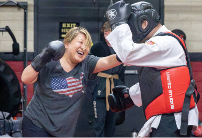 A middle-aged woman is punching a larger man in full-body protective gear as she participates in a women's self defense class.