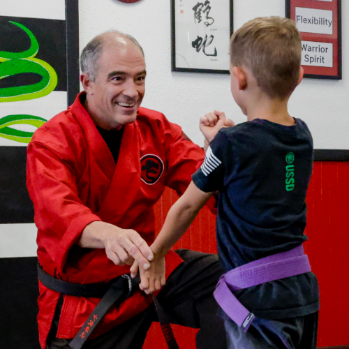 Chief Instructor Sensei Mike is on his knee and smiling at a young purple belt student while gently guiding him through a series of new block positions.