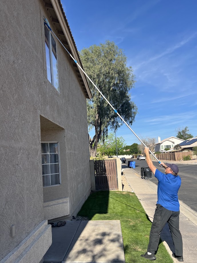 Person wearing a blue shirt and cap using a long-handled tool to clean the exterior window of a beige house on a sunny day.