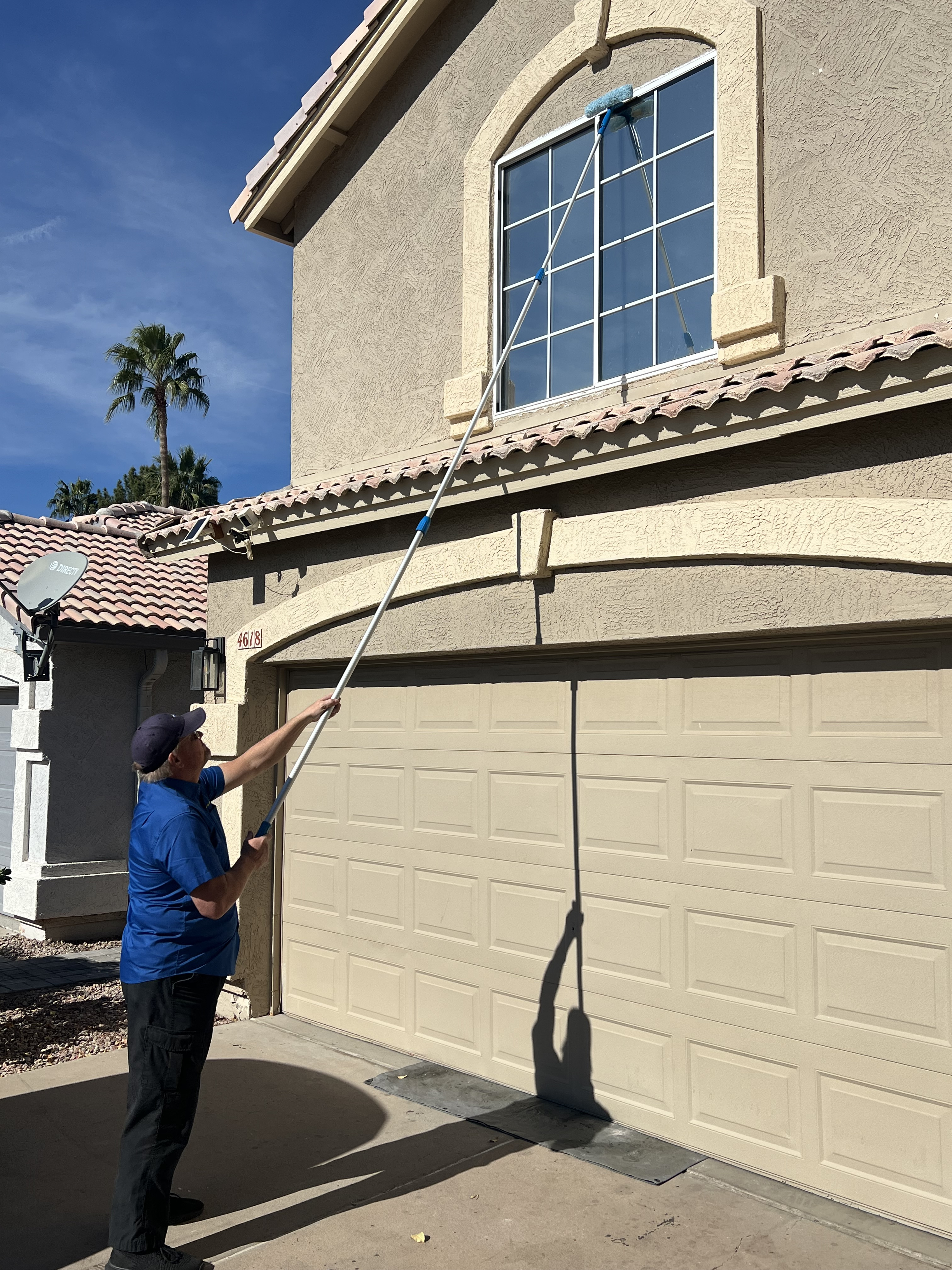 Man in blue shirt and black cap cleaning a second-story window of a beige house using a long pole with a window cleaning brush.