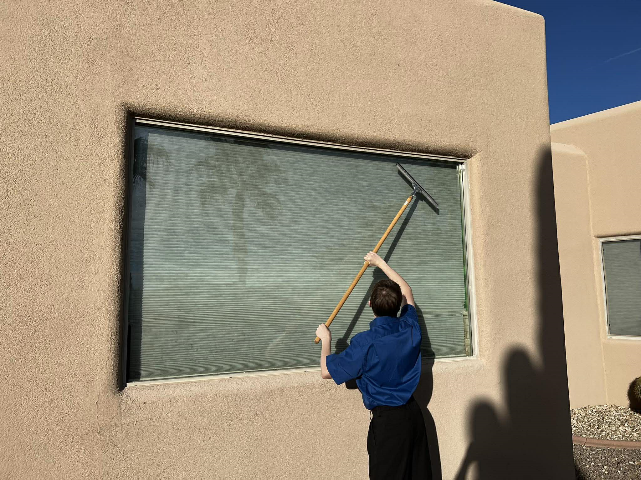 Person in a blue shirt using a long-handled squeegee to clean a large outdoor window on a beige stucco building.
