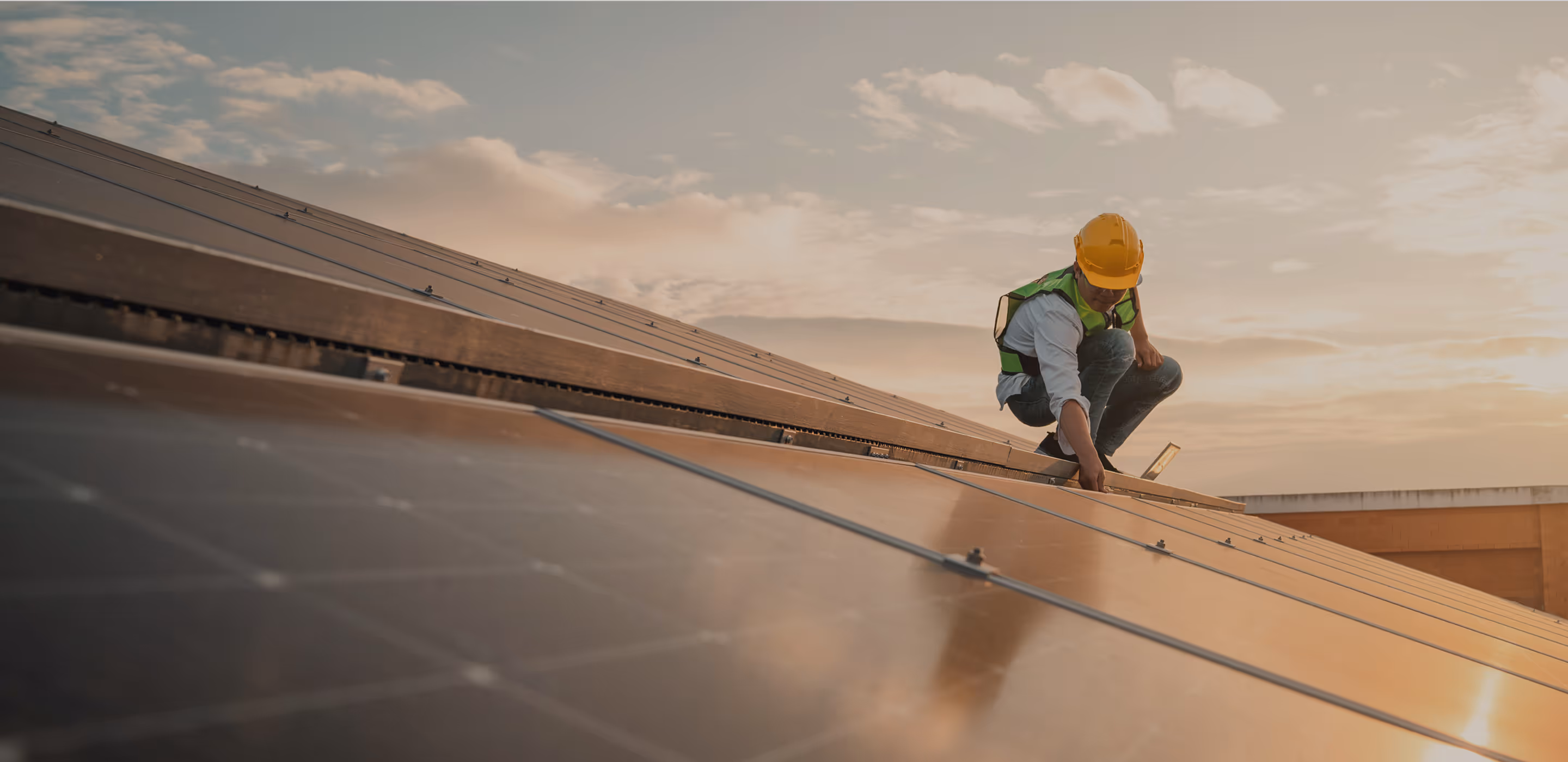 Solar technician in a yellow safety helmet and reflective vest inspects solar panels on a slanted roof during sunset, with warm light reflecting off the panels.