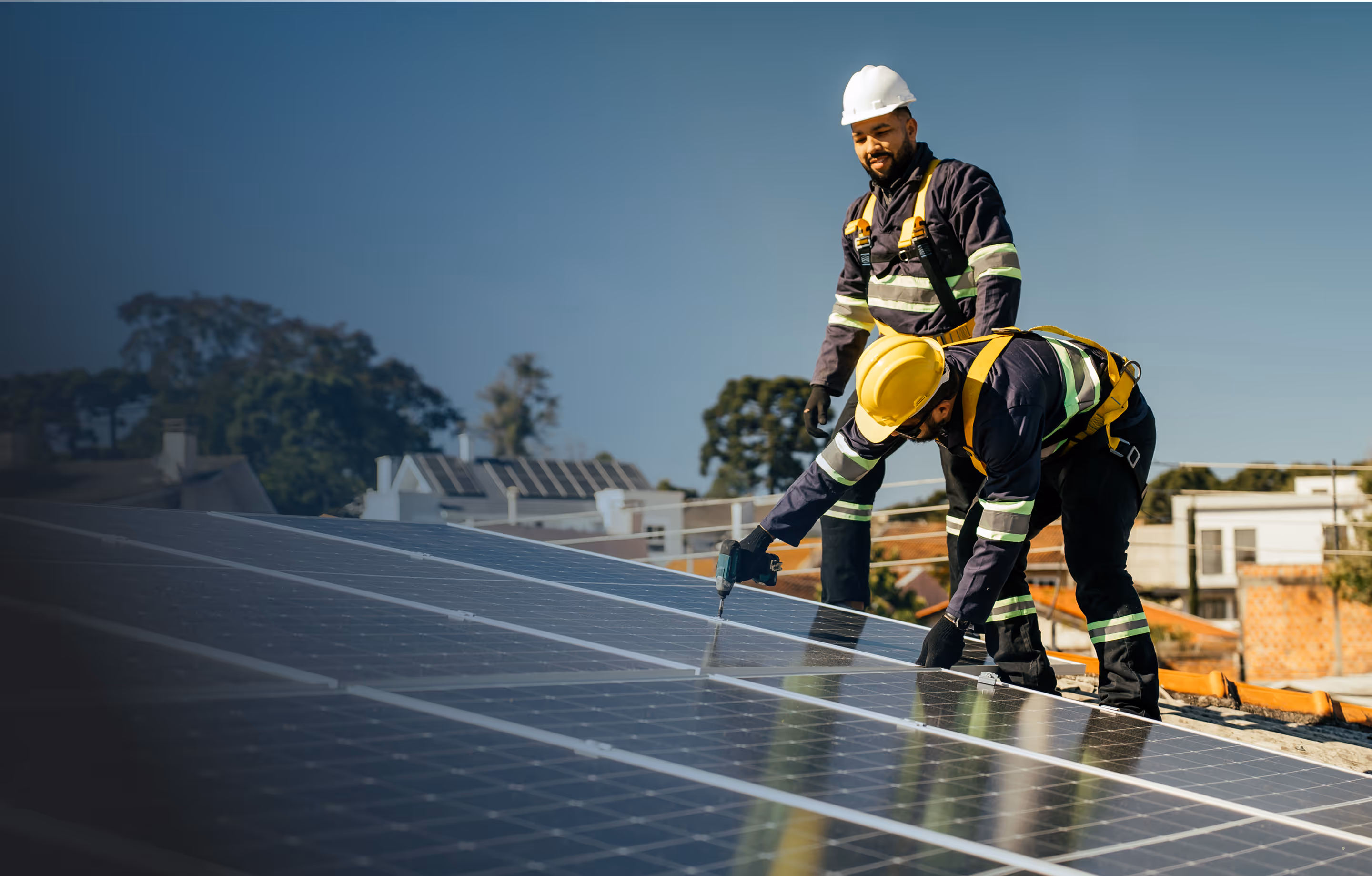 Two construction workers in safety gear install solar panels on a residential rooftop under a clear blue sky; one uses a power drill while the other observes. Homes with more solar panels and trees are visible in the background.