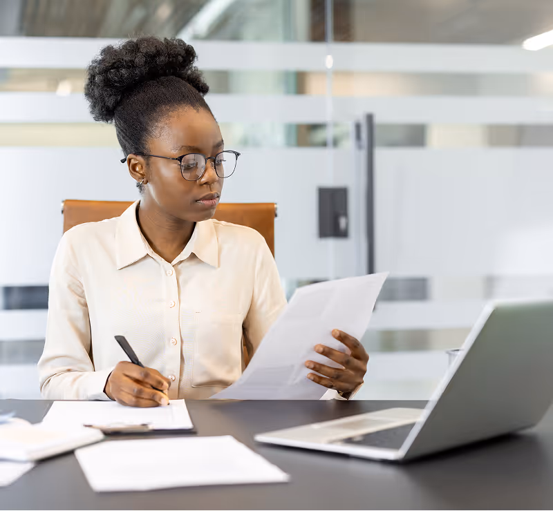 Woman wearing glasses sits at a modern desk, reviewing a printed document while writing notes beside an open laptop in a glass-walled office.